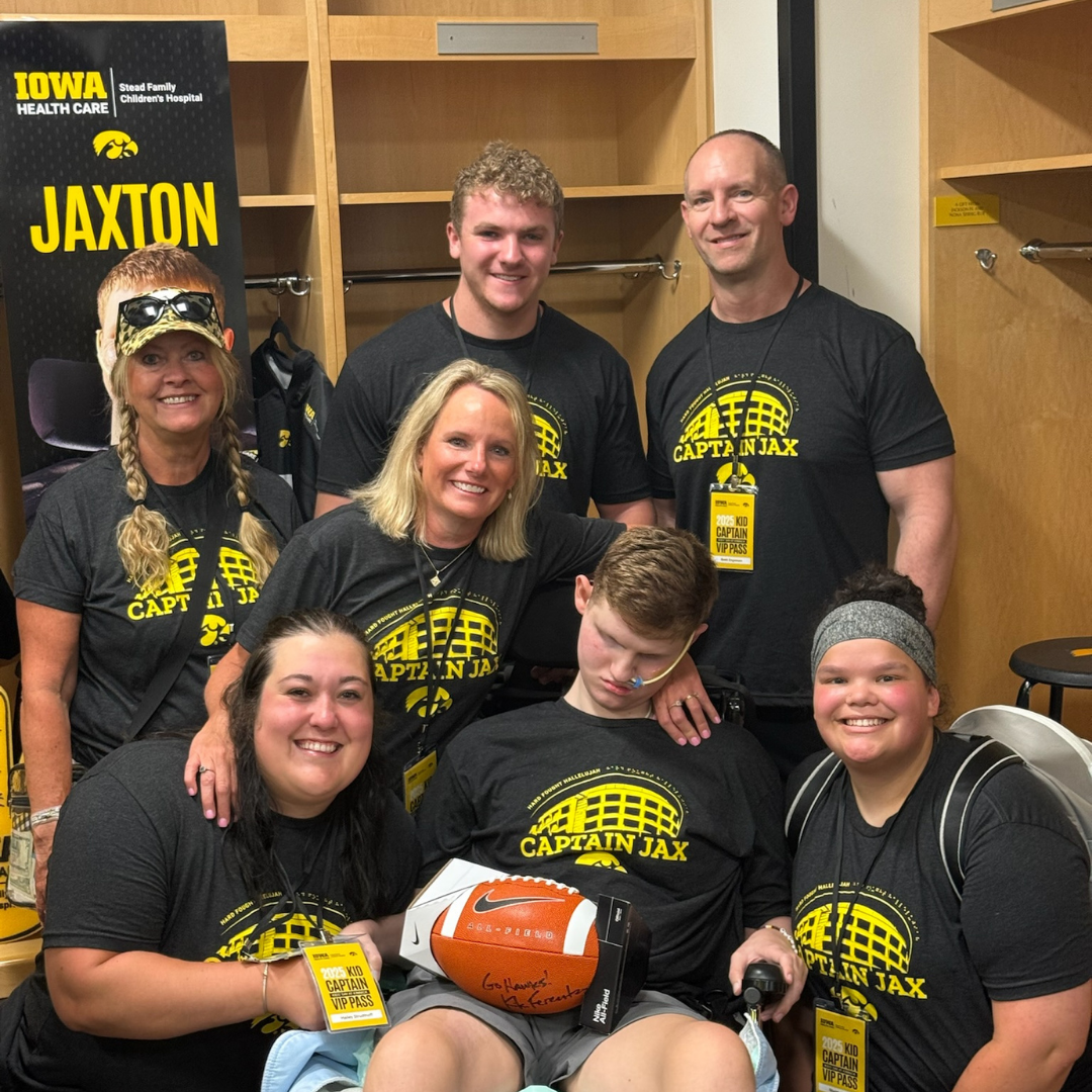 A young boy honored as a University of Iowa Kid Captain sits in a wheelchair holding a signed football, surrounded by smiling family and supporters all wearing matching black ‘Team Captain Jax’ t-shirts inside a locker room.